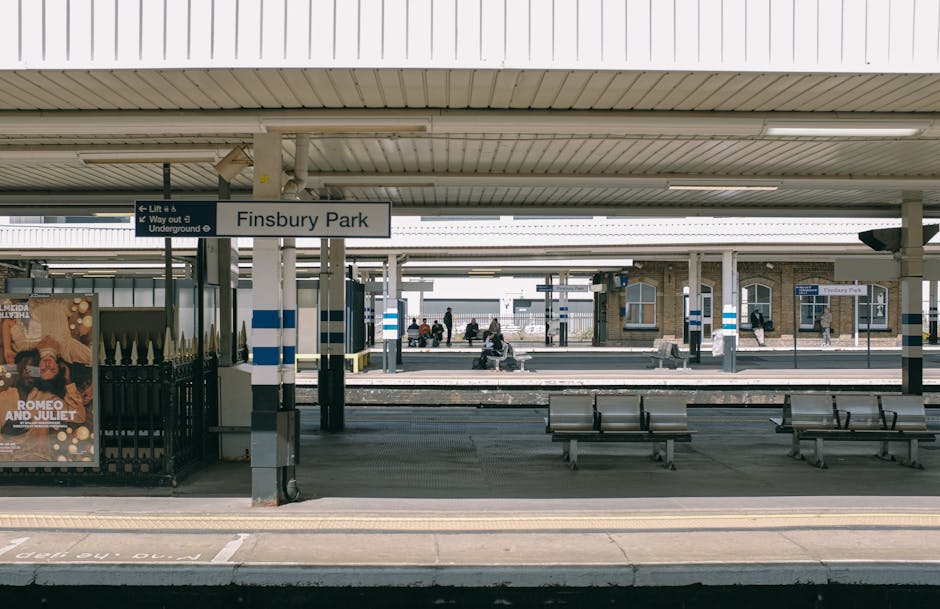 The image shows the platform of Finsbury Park train station with a sign indicating the station name, located in London’s NW3 area. The platform is covered by a roof supported by metal poles, with a row of benches for waiting passengers. Several people are seated on the benches or walking along the platform. On the left side, there is a black metal fence and a poster advertising a theatrical production of 'Romeo and Juliet.' The platform surface features tactile paving for visually impaired travelers and safety markings, with a yellow line near the edge indicating the safe distance from the track. In the background, additional platforms and railway tracks extend into the distance, with more passengers visible waiting or walking. Natural daylight illuminates the scene, and the environment emphasizes a typical urban train station setting suitable for home relocation or moving logistics, as handled by Belsize Park Removals.
