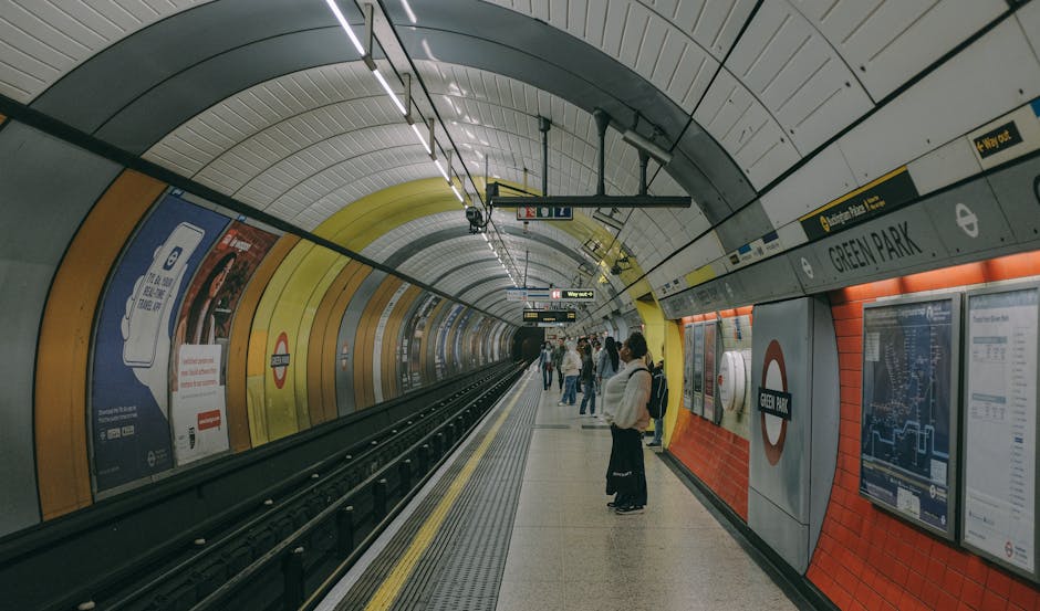 Inside the Green Park London Underground station showing the curved, tiled walls with orange and yellow acoustic panels, illuminated by overhead lighting fixtures. The platform has a tactile yellow line near the edge for safety, and several passengers are waiting, some standing near information posters and electronic station maps. The station signage indicates 'Green Park' and includes the London Underground roundel logo. The tunnel extends into the darkness ahead, with train tracks running along the middle of the platform. This setting is relevant for a house removals service, illustrating the transport environment involved in packing and moving furniture during a home relocation process, supported by Belsize Park Removals' professional services.