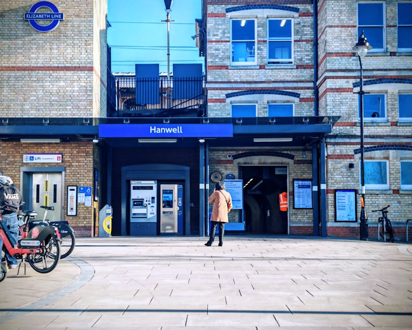The image shows the platform of Finsbury Park train station with a sign indicating the station name, located in London’s NW3 area. The platform is covered by a roof supported by metal poles, with a row of benches for waiting passengers. Several people are seated on the benches or walking along the platform. On the left side, there is a black metal fence and a poster advertising a theatrical production of 'Romeo and Juliet.' The platform surface features tactile paving for visually impaired travelers and safety markings, with a yellow line near the edge indicating the safe distance from the track. In the background, additional platforms and railway tracks extend into the distance, with more passengers visible waiting or walking. Natural daylight illuminates the scene, and the environment emphasizes a typical urban train station setting suitable for home relocation or moving logistics, as handled by Belsize Park Removals.
