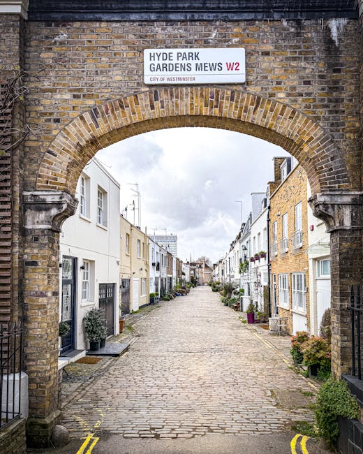 A narrow residential street in Belsize Park with a steep incline, featuring mostly brick and white-painted houses. The pavement is lined with potted plants and flower boxes attached to iron railings a