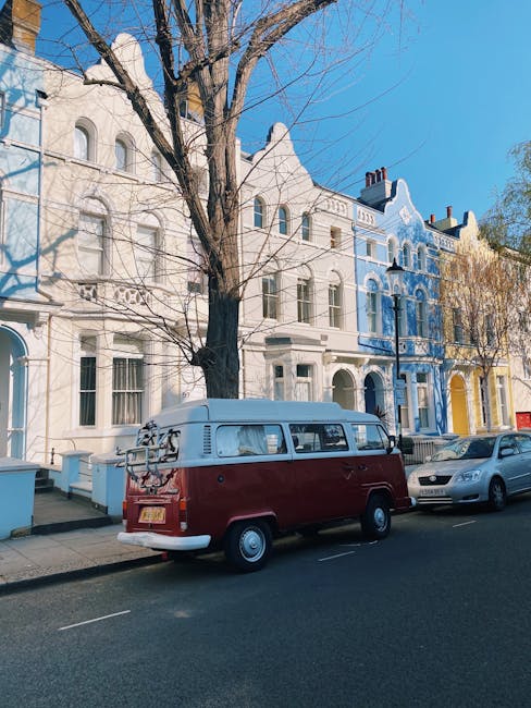 A vintage red and white Volkswagen van, parked on a residential street in Belsize Park, London, with a row of colorful Victorian-style terraced houses behind it. The van has a bicycle rack mounted on the rear and is positioned on the pavement adjacent to a silver car. The street scene is illuminated by natural daylight, with a large leafless tree standing alongside the houses. This image depicts a scene relevant to home relocation and furniture transport processes that might be associated with house removals services provided by Belsize Park Removals, including the transport of boxes and furniture during packing and loading for a house move on Haverstock Hill.
