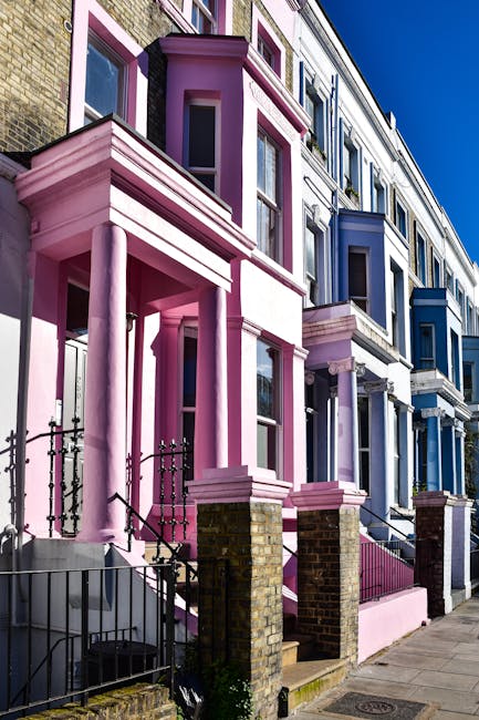 An exterior view of a row of colorful Victorian terraced houses featuring prominent bay windows and decorative cornices, with facades painted in various shades including pink, purple, and blue. The houses have front steps leading to entrances, some with railings, and are positioned along a paved sidewalk. The image captures a bright, clear day with sunlight casting shadows on the buildings. This scene reflects a typical residential street where house removals and home relocations might involve careful packing, furniture transport, and loading processes to ensure safe moving of belongings, as handled by companies such as Belsize Park Removals.
